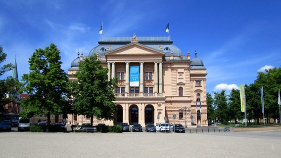 Das Gebäude des Mecklenburgischen Staatstheaters Schwerin bei Sonnenschein mit wehenden Fahnen, Bäumen und parkenden Autos © NDR Foto: Axel Seitz