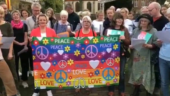 Etwa zwanzig Menschen stehen hinter einem bunten Plakat, das mit "Peace"- und "Love"-Schriftzügen sowie Herzen, Blumen und Peace-Zeichen bemalt ist © NDR 