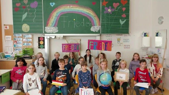 Schülerinnen und Schüler einer Grundschulklasse sitzen vor einer bunt bemalten Tafel und halten selbstgebastelte Schilder mit der Aufschrift "Frieden" und "3a" in den Händen © NDR 