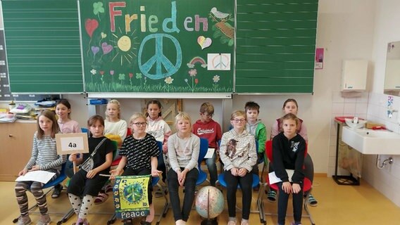 Schülerinnen und Schüler einer Grundschulklasse sitzen vor einer bunt bemalten Tafel und halten selbstgebastelte Schilder mit der Aufschrift "Frieden" und "4a" in den Händen © NDR 