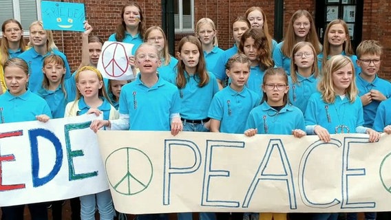 Etwa zwanzig Kinder in türkisen Poloshirts halten beim Singen ein Plakat mit der Aufschrift "Peace" © NDR 