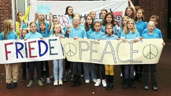 Etwa zwanzig Kinder in türkisen Poloshirts halten beim Singen Plakate mit den Aufschriften "Friede" und "Peace" © NDR 
