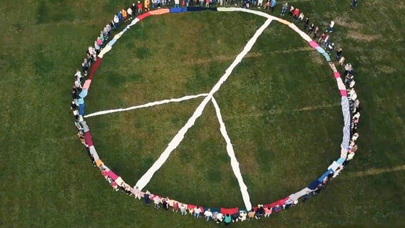 Eine große Menschengruppe steht auf einer Wiese im Kreis um ein aus Stoff gelegtes Peace-Zeichen © NDR 