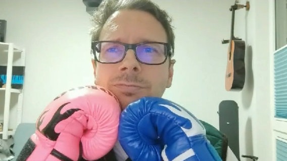 Ein Mann mit Brille und je einem rosa und blauen Boxhandschuh schaut in die Kamera © NDR 