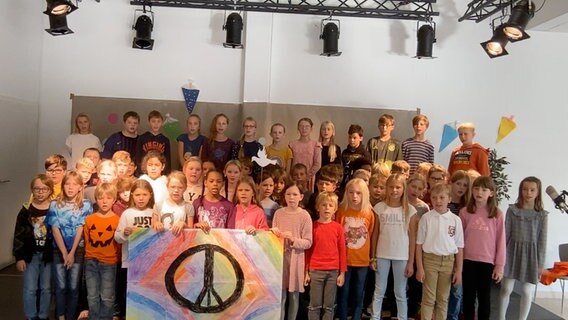 Etwa fünfzig Kinder im Grundschulalter stehen auf einer Bühne. Sie halten ein selbstgemaltes Plakat mit einem Peace-Zeichen © NDR 