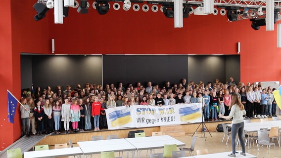 Etwa fünfzig Kinder stehen singend auf einer Bühne. Sie halten ein Plakat mit der Aufschrift "Stop War. Wir gegen Krieg!" sowie eine EU- und eine Ukraine-Flagge © NDR 