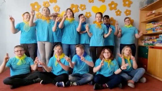 Etwa fünfzehn Kinder mit türkisen T-Shirts und gelben Halstüchern singen zusammen in einem Klassenraum © NDR 