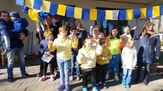 Etwa zehn Kinder stehen mit ein paar Erwachsenen unter einer blau-gelben Girlande und singen © NDR 