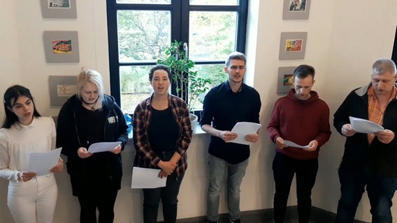Sechs Menschen stehen in einer Reihe, halten Papierseiten in der Hand und singen © NDR 
