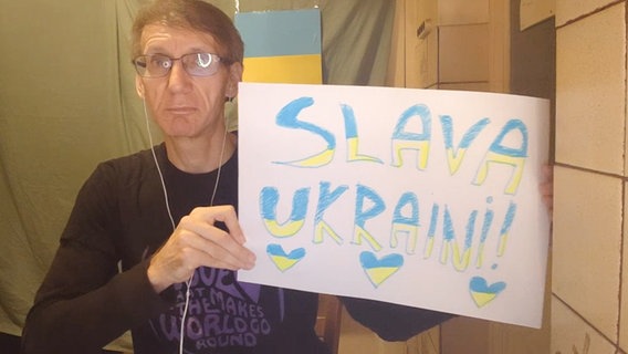 Ein blonder Mann mit Brille hält ein selbstgemaltes Schild mit der Aufschrift "Slava Ukraini!" in die Kamera © NDR 