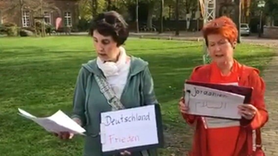 Zwei Frauen stehen singend auf einer Wiese und halten Blätter mit den Worten "Deutschland", "Frieden" und "Jordanien" in den Händen © NDR 