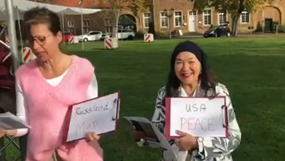 Zwei Frauen stehen singend auf einer Wiese und halten Blätter mit den Worten "Russland", "мир", "USA" und "Peace" in den Händen © NDR 