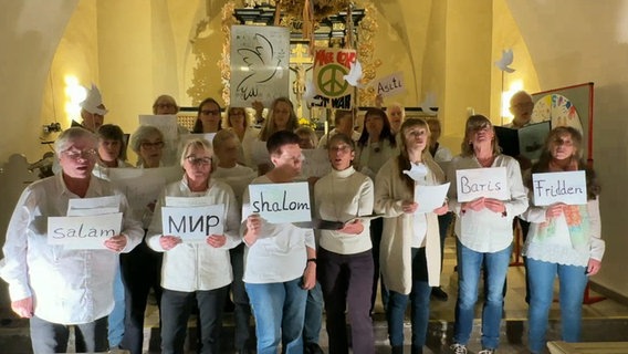 Etwa zwanzig Menschen in heller Kleidung stehen singend in einer Kirche und halten Blätter mit dem Wort "Frieden" in verschiedenen Sprachen © NDR 