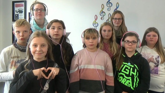 Neun Kinder und Jugendliche mit Kopfhörern stehen in drei Reihen, an der Wand hinter ihnen sind Notenschlüssel zu sehen © NDR 