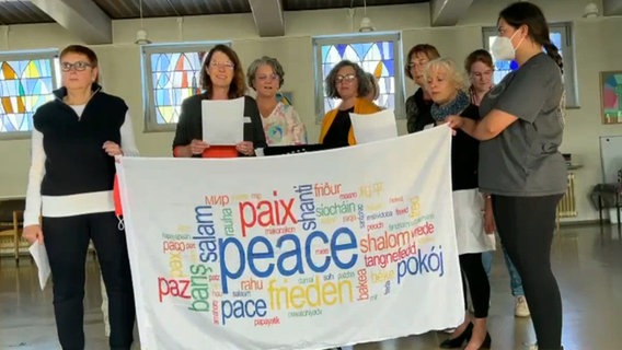 Etwa zehn Frauen singen und halten eine weiße Fahne, in der auf verschiedenen Sprachen das Wort "Frieden" steht © NDR 