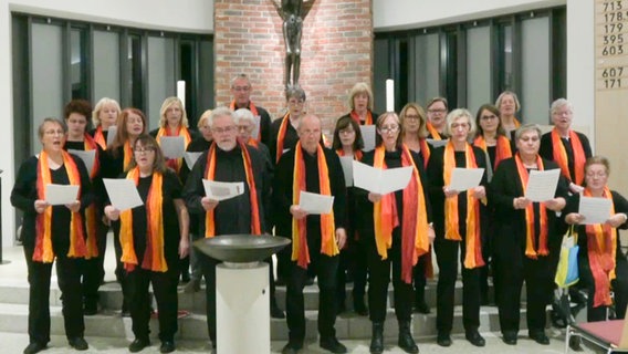 Ein gemischter Chor in schwarzer Kleidung und orangenen Schals steht vor einem Altar © NDR 