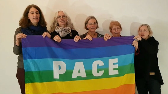 Fünf Frauen stehen singend nebeneinander und halten eine Regenbogenfahne mit der Aufschrift "Pace" © NDR 