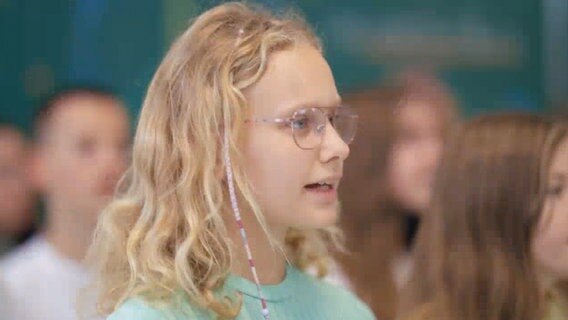 Ein Mädchen mit blonden Locken und Brille singt, im Hintergrund sieht man verschwommen andere Kinder und Jugendliche © NDR 