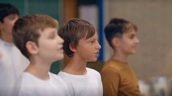 Drei Jungen stehen in einer Reihe und singen, hinter ihnen sieht man verschwommen einen weireren Jungen © NDR 