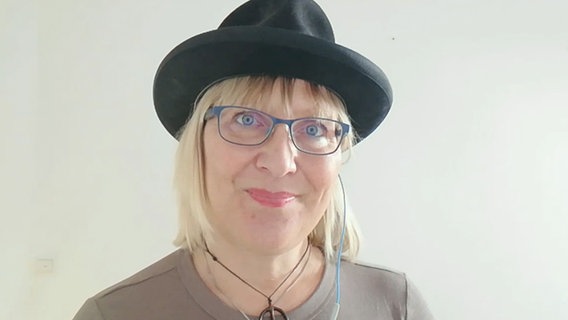 Eine blonde Frau mit Brille und schwarzem Hut lächelt in die Kamera © NDR 