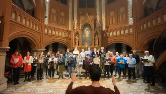 Ein gemischter Chor mit etwa dreißig Mitgliedern steht vor einem Altar und singt © NDR 