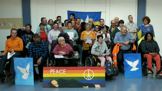 Etwa dreißig Menschen stehen und sitzen in drei Reihen, vor ihnen sind Bilder mit Friedenstauben aufhetsellt, einem Regenbogen und einem "Peace"-Schriftzug, hinter ihnen hängt eine Ukraine-Flagge © NDR 