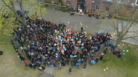 Eine große Anzahl von Jugendlichen steht mit gelben und blauen Luftballons auf einem Schulhof © NDR 