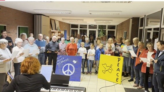 Etwa vierzig Menschen aller Altersgruppen stehen in einem Halbkreis und singen, vor ihnen stehen ein blaues Bild mit der Aufschrift "give peace a chance" und ein gelbes Bild mit einer Friedenstaube © NDR 