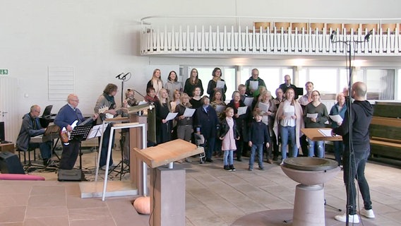 Ein Chor mit etwa zwanzig Mitgliedern singt in einer Kirche und wird von einem Klavier, einer E-Gitarre und einem E-Bass begleitet © NDR 