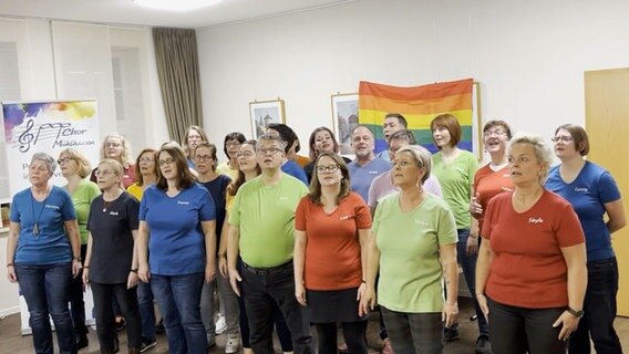 Etwa fünfundzwanzig Menschen stehen zusammen und singen, hinter ihnen hängt eine Regenbogenfahne und ein Banner mit der Aufschrift "Chor Mühlhausen" © NDR 
