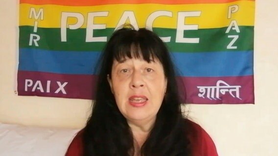 Eine Frau mit schwarzen Haaren sitzt vor einer Regenbogenfahne mit der Aufschrift "Peace" und singt © NDR 