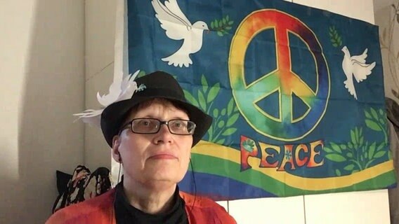 Eine Frau mit schwarzem Hut und Brille sitzt neben einem Tuch mit Friedenstauben und einem Peace-Zeichen © NDR 