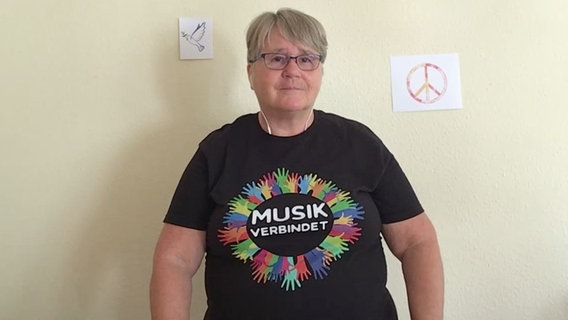 Eine Frau mit kurzen, grauen Haaren und Brille schaut in die Kamera. Sie trägt ein T-Shirt mit der Aufschrift "Musik verbindets" © NDR 