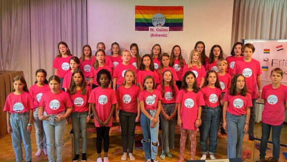 Etwa dreißig Kinder in pinken T-Shirts stehen vor einer Regenbogenfahne, unter der ein Schild mit der Aufschrift "St. Gallen (Schweiz)" hängt © NDR 