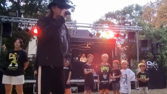 Sechs Kinder mit schwarzen Udo-Shirts und ein Erwachsener im Udo-Lindenberg-Style mit Mikrofon singen auf einer Open Air-Bühne. © NDR 