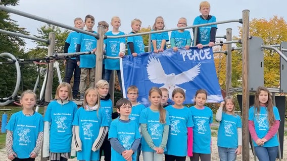 Knapp zwanzig Kinder in bedruckten türkisfarbenen T-Shirts stehen vor und auf einem Klettergerüst und blicken in die Kamera. Die Kinder auf dem Gerüst halten eine große blaue Flagge mit einer weißen Friedenstaube und dem Wort "Peace". © NDR 