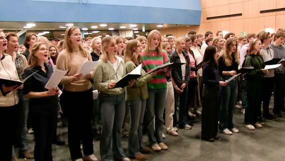 Eine Gruppe von mehr als fünfzig Jugendliche steht mit Mappen und Zetteln in der Hand in einer Art Foyer und singt. © NDR 