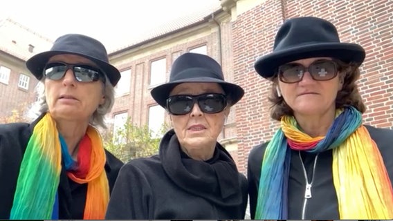 Drei schwarz gekleidete Frauen mit schwarzen Sonnenbrillen und schwarzen Hüten stehen nebeneinander und blicken in die Kamera. Die mittlere trägt einen schwarzen Schal, die beiden anderen jeweils einen regenbogenfarbenen. Im Hintergrund ist ein Backsteingebäude zu erkennen. © NDR 