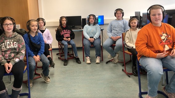 Acht Kinder mit Kopfhörern sitzen in einem Raum mit Computern auf Stühlen und singen mit Blick zur Kamera. © NDR 