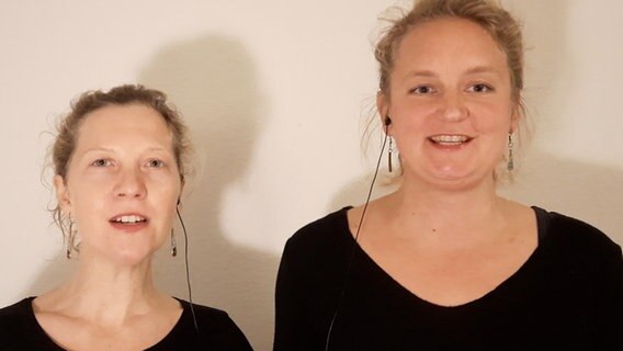 Zwei schwarz gekleidete, blonde Frauen mit Zopf blicken singend in die Kamera. Jede benutz je einen Teil eines In-ear-Kopfhörers. © NDR 