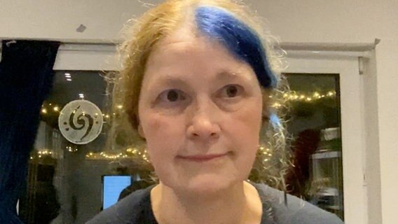 Nahaufnahme einer Frau mit in der Mitte gescheiteltem blonden Haar, das auf ihrer linken Seite blau gefärbt ist. Auf dem Fenster im Hintergrund ist ein Ornament aus zwei verschlungenen Bassschlüsseln zu erkennen. © NDR 