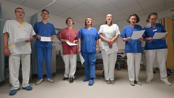 Sieben Erwachsene in blauen, weißen bzw. roten Kasacks mit Zetteln in der Hand haben sich in einem Raum aufgestellt und singen. Im Hintergrund befinden sich ein Krankenhausbett und medizinische Gerätschaften. © NDR 