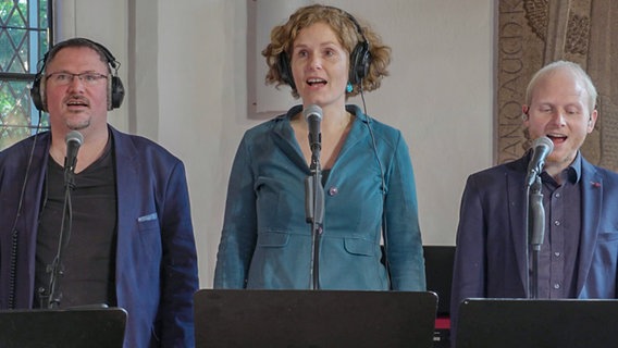 Eine Frau und zwei Männer in blauen Sakkos und mit Kopfhörern singen jeweils in ihr Standmikrofon. Im Hintergrund sind ein Kirchenfenster und eine alte Grabplatte zu erkennen. © NDR 