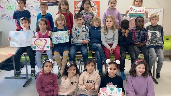 Zwanzig Kinder sitzen und stehen in drei Reihen, halten selbstgemalte Bilder mit den Worten "Frieden" hoch und singen. © NDR 