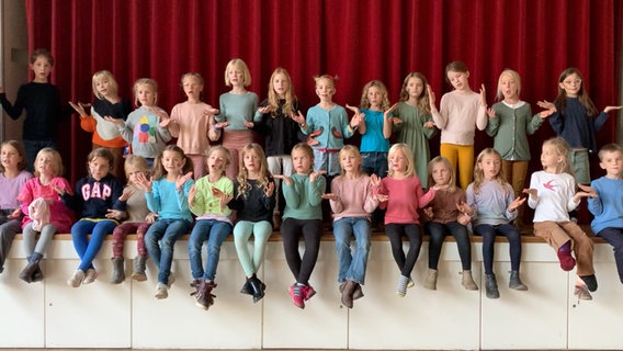 26 Kinder sitzen und stehen in zwei Reihen auf einer Bühne vor einem geschlossenen roten Vorhang. Sie halten ihre Handflächen seitlich auf und singen. © NDR 