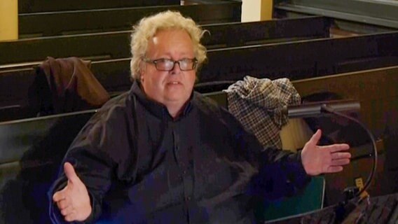 Ein Mann mit kurzen, grauen Locken und schwarzer Brille sitzt mit ausgebreiteten Armen in der vordersten Kirchenbank, vor sich ein Notenpult mit Lampe, auf den hinteren Bänken sind Jacken verstreut. © NDR 