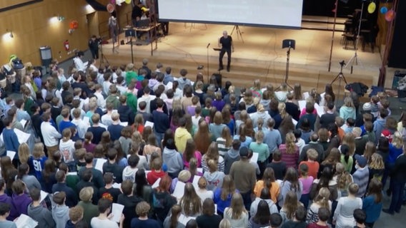 Eine große Anzahl an Schülern hat sich in einer Aula zum Singen versammelt und wird dort von einem Mann dirigiert © NDR 