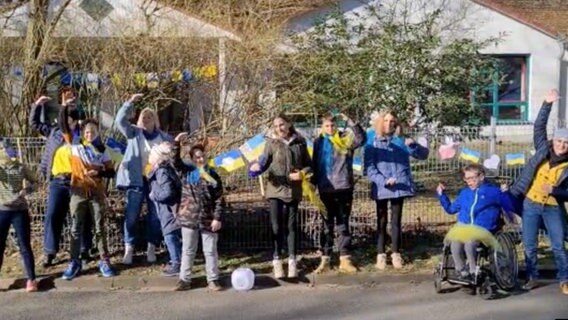 Etwa zehn Kinder singen zusammen auf dem Schulhof. Einige sind blau-gelb angezogen, am Zaun hängt eine Girlande mit kleinen Ukraine-Fahnen © NDR 