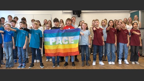 Über dreißig Kinder, größtenteils in blau-grünen oder roten T-Shirts haben sich in mehreren Reihen aufgestellt, sie singen und machen eine nach vorn weisende Geste. Vorn in der Mitte halten einige eine große Flagge in Regenbogenfarben mit weißem Schriftzug "Pace". © NDR 