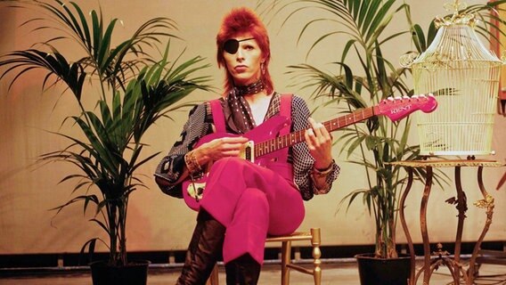 David Bowie sitzt auf einem Stuhl und hält eine E-Gitarre in der Hand, daneben ein leerer Vogelkäfig. © picture alliance / Photoshot 